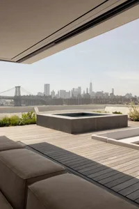 A modern rooftop terrace features a wooden deck, a concrete fireplace, and plants with a scenic view of the city skyline.