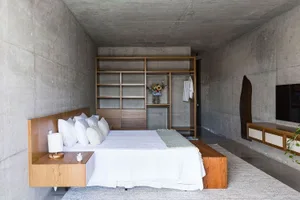 A minimalist bedroom with a concrete wall and wooden furniture, featuring a bed with white linens and a built-in wardrobe.