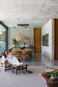 A modern living room with concrete walls, wooden seating, and a large wicker pendant light hanging above. A person is relaxin