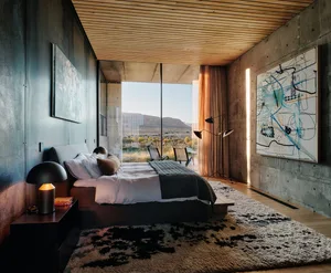 Modern bedroom with concrete walls, wooden flooring, and large glass windows overlooking a natural landscape.