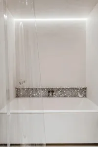 Modern minimalist bathroom featuring a white bathtub with a textured terrazzo backsplash and illuminated lighting.