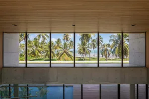 Large glass window overlooking a tropical beach with palm trees and blue sky.