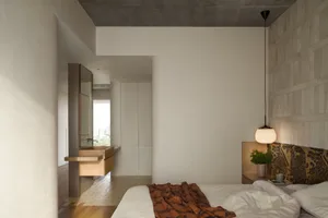 A minimalist bedroom with concrete and wooden elements, featuring a bed with a blanket and a hanging lamp.