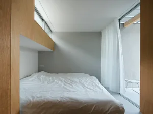 A minimalist bedroom with a white bed, white curtains, and light wooden walls.