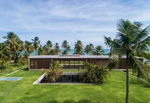 A modern house with a concrete exterior surrounded by lush greenery and palm trees, overlooking the ocean.