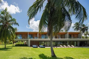 Modern house with concrete exterior and large glass windows, surrounded by greenery and palm trees.