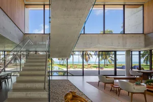 Modern living room with concrete walls and floor, large windows overlooking the sea, and a glass staircase.