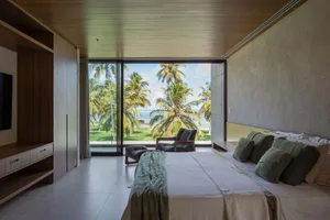 Modern minimalist bedroom featuring a bed with wooden headboard and floor-to-ceiling glass window overlooking palm trees and 