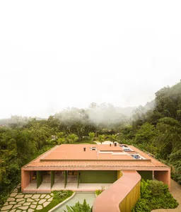 A modern residential building with terracotta roof and concrete walls surrounded by lush green vegetation.
