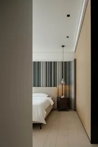 A minimalist bedroom featuring a white bed with a striped wall and a side table with a lamp.