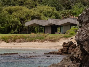 Modern house with geometric roofline on a sandy beach with lush greenery in the background.