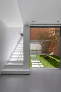 A minimalist interior featuring a white staircase and a glass door leading to a garden with a tree and stone pathways.