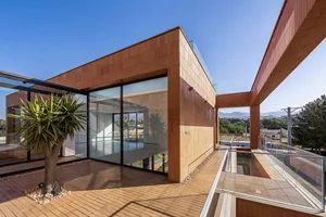 A modern residential building with a wooden pool and glass walls, featuring a terrace and a palm tree.