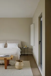 A minimalist bedroom with a neutral color palette, featuring a bed, side table with a bottle, and woven basket on a light-col