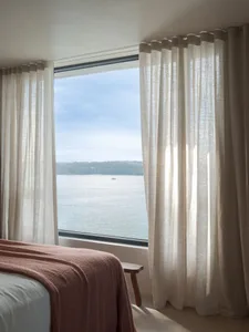 A bedroom with a bed covered in a reddish-brown blanket, floor-to-ceiling curtains with sheer white fabric, and a view of a c
