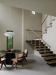 Modern residential dining space with round wooden table, rattan back chairs, minimalist staircase, and large window.