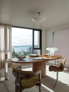 A modern residential office space featuring a wood desk and chairs, with books and a lamp on the desk, and a large window ove