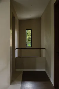 A corridor with neutral-colored walls and floors, featuring a window with a view of greenery and a wooden railing.