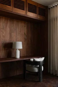 A simple home office featuring a dark wood desk, a black chair with a white cushion, a white table lamp, and wood-paneled wal