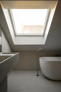 A minimalist bathroom featuring concrete walls and a freestanding tub with a skylight above.