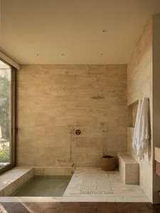 A minimalist bathroom with stone walls, a walk-in shower, and a stone bathtub. The space is illuminated by natural light from