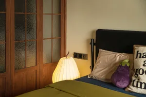 Minimalist bedroom featuring a purple pillow, green bedspread, and stylish yellow pear-shaped lamp on a bed with a black upho