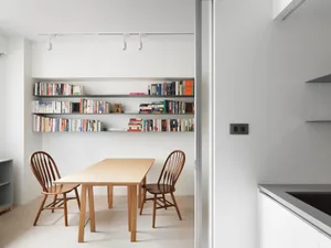 Minimalist dining room featuring a wooden table, wooden chairs, and a metal bookshelf filled with books.