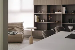 A minimalist living room featuring a beige leather sofa, a white round side table, and a wooden shelving unit with books and 