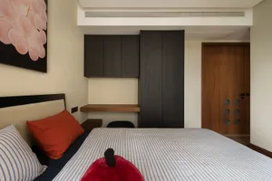 A modern bedroom featuring light-colored wooden furniture, a blue and white striped bedspread, and red and orange accent pill