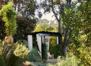 A modern pavilion with white curtains and a wooden chair surrounded by lush greenery and trees.