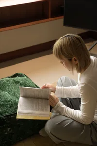 A girl sitting on a green couch, reading a book with a pen in her hand.
