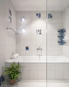 A minimalist bathroom with white ceramic tiles, blue accents, a white bathtub, and a plant.