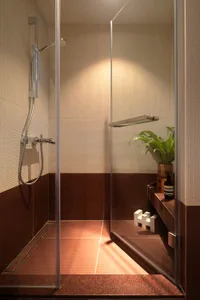A minimalist bathroom featuring red tiles on the lower half of the walls, with a glass shower enclosure and a potted plant.