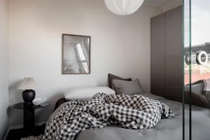 A minimalist bedroom featuring a bed with a checkered blanket, pillows, and a lamp on a bedside table.