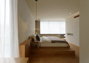 A minimalist bedroom featuring a wooden platform bed with white linens, a single chair, and large windows allowing natural li