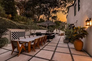 A modern backyard patio features a concrete and glass dining area with seating and an umbrella for shade.