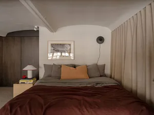 A modern bedroom with mixed materials including a bed with a duvet, pillows, and a headboard. The room features a desk lamp a