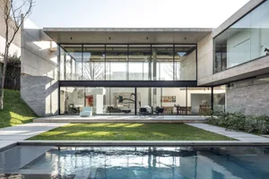 A modern residential home with concrete walls and glass walls and doors, featuring a swimming pool and a large green lawn.
