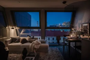 A cozy modern living room with a city view at dusk, featuring soft furnishings and natural light.