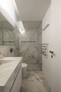 Modern minimalist bathroom with marble walls and floor, featuring a walk-in shower with glass door, a toilet, and a vanity si