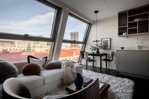 A modern living room with a view of the city, featuring a white sofa with pillows and a blanket, a round cushion, a white cof