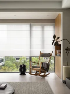 A modern minimalist living room with a large window covered by white blinds, featuring a wooden rocking chair with a cushion