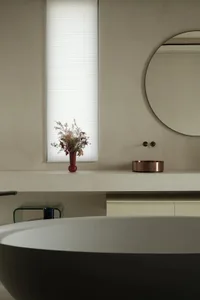 Modern minimalist bathroom interior with concrete walls, a round mirror, a vase with dried flowers, and a large window.