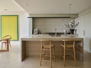 A modern, minimalist residential kitchen with wood stools, a stone countertop, and a large yellow painting on the wall.