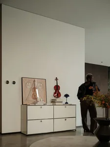 一間現代客廳，擺放白色家具、提琴，以及牆上裱框的提琴素描。室內站著一位人物。