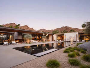 Modern minimalist residential exterior with a swimming pool and fire pit, surrounded by desert landscape and mountains in the