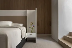 A minimalist bedroom featuring a bed with white bedding, a wooden headboard, and a nightstand with a vase of flowers. The roo