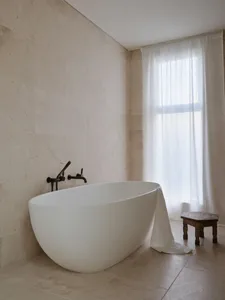 A minimalist bathroom featuring a freestanding white marble bathtub, black fixtures, and a large window with sheer curtains a