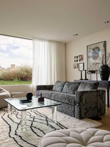 A modern living room featuring a patterned sofa, white curtains, and a glass coffee table.