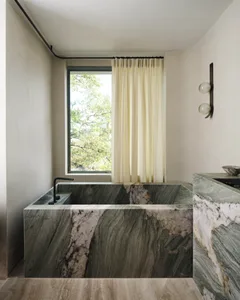 Modern marble bathroom with green and white marble bathtub and floor-to-ceiling curtains by a window.